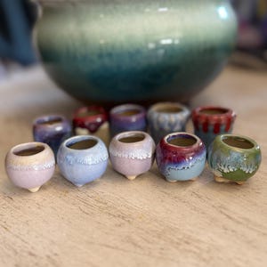 May include: A collection of small, handcrafted ceramic pots in various colors, including blue, purple, and green, are arranged on a wooden surface. A larger, two-toned green and blue ceramic pot sits in the background, with a green plant inside.