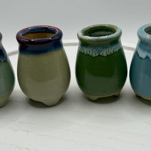 May include: Four small ceramic pots with rounded bodies and three short feet. Each pot features a unique reactive glaze finish in shades of green, blue, and purple, with a smooth, glossy texture on the upper sections and a matte base.