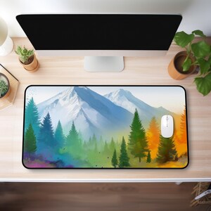 Pode incluir: Um tapete de rato retangular com uma paisagem em aquarela de uma cordilheira e árvores perenes. A paleta de cores inclui tons de azul, verde e laranja. Um rato de computador branco está sobre o tapete.