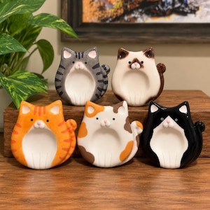 May include: Five ceramic cat-shaped trinket dishes arranged in two rows. The top row features a gray tabby and a cream-colored cat with brown points. The bottom row includes an orange tabby, a white calico, and a black and white cat.