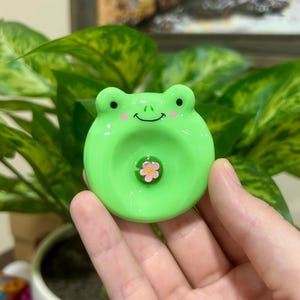 May include: A round, light green ceramic frog-shaped object with a smooth, glossy finish. The frog features two small black eyes, a simple black curved mouth, and two pink circular cheek accents. A small pink flower is centered on the frog's belly.