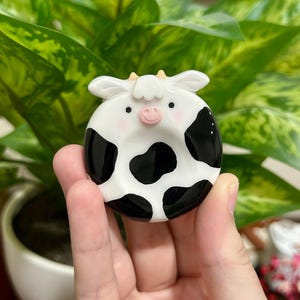 May include: A round, ceramic-style trinket dish shaped like a cow, featuring a white base with black spots, a small pink snout, two tiny horns, and rosy cheeks. The surface is smooth and glossy, with a central indentation for holding small items.