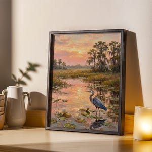 May include: Framed landscape painting featuring a serene marsh scene with a great blue heron. The artwork showcases a sunset with warm hues of orange and pink, reflecting in the water. The heron stands among lily pads, adding a touch of wildlife to the tranquil setting.