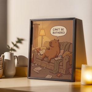 May include: Framed art print depicting a cartoon capybara relaxing on a sofa, with the text "CAN'T BE BOTHERED" in a speech bubble. The artwork uses warm colours, with books and crumpled paper scattered around.