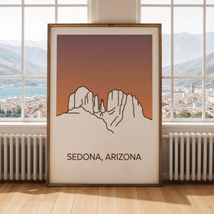 May include: A framed print of a minimalist line drawing depicting the Sedona, Arizona landscape. The artwork features a gradient orange and brown sky, with the text "SEDONA, ARIZONA" at the bottom. The print is displayed in a room with a window and a radiator.