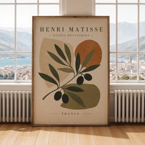 May include: A framed art print featuring a botanical illustration in a minimalist style. The print includes a branch with olives, abstract shapes in earth tones, and the text "HENRI MATISSE - ÉTUDES BOTANIQUES - FRANCE". The artwork is displayed in a room with a window.