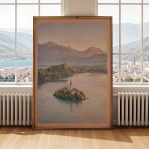 Puede incluir: Una pintura de paisaje enmarcada representa una serena escena lacustre con una pequeña iglesia en una isla. La obra de arte presenta colores suaves y apagados de marrón, verde y azul, con montañas al fondo. La pintura se exhibe en una habitación con ventana y suelo de madera.