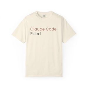 Può includere: T-shirt color crema con la scritta "Claude Code Pilled" in un gradiente marrone e rosso. La maglietta ha un girocollo e maniche corte, adatta per l'abbigliamento casual.