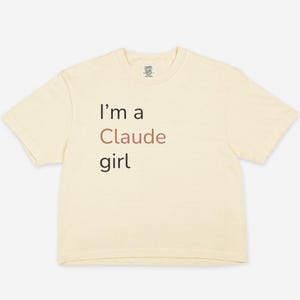 Könnte beinhalten: Cremefarbenes T-Shirt mit dem Text "I'm a Claude girl" in schwarzer und brauner Schrift. Das Shirt hat einen Rundhalsausschnitt und kurze Ärmel. Das Comfort Colors-Logo ist oben in der Mitte des Shirts sichtbar.