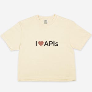 Può includere: T-shirt color crema con la scritta "I ❤ APIs" in nero e un simbolo a cuore. La maglietta ha girocollo e maniche corte. Il tessuto sembra morbido e confortevole.