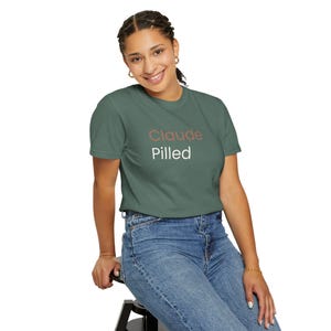 May include: A green t-shirt featuring the text "Claude Pilled". The word "Claude" is in a muted red, and "Pilled" is in white. The shirt is worn with blue jeans. The person is smiling.