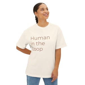 Peut inclure: T-shirt crème avec l'inscription "Human in the Loop" en marron rougeâtre. Le t-shirt a des manches courtes et un col rond. Le modèle porte un jean bleu.