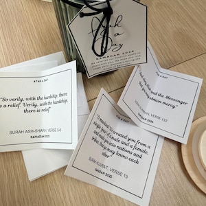 Puede incluir: Tarjetas blancas y negras de Ayah a Day con versos islámicos y el texto "Ramadán 2025". Las tarjetas están en un recipiente de vidrio transparente con una etiqueta negra. Las tarjetas están sobre una superficie de madera clara.