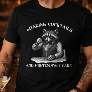 Könnte beinhalten: Ein schwarzes Kurzarm-T-Shirt mit einer Grafik eines Waschbären, der als Barkeeper in Weste und Fliege gekleidet ist und einen Cocktail rührt. Der Text darüber lautet SHAKING COCKTAILS und darunter AND PRETENDING I CARE in weißen Buchstaben.
