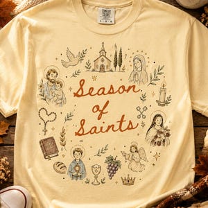 May include: A pale yellow short-sleeve t-shirt featuring a circular graphic print with the text Season of Saints in the center. The design includes illustrations of a church, a dove, religious figures, a rosary, a bible, bread, and grapes.