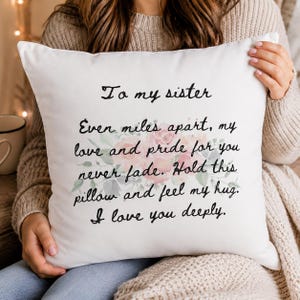 Op de afbeelding: Een vierkant wit sierkussen met een bloemrijk aquarelontwerp van roze rozen en groene bladeren in het midden. Het kussen is bedrukt met zwarte cursieve tekst die luidt: To my sister, Even miles apart, my love and pride for you never fade. Hold this pillow and feel my hug. I love you deeply.