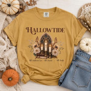 Può includere: Una t-shirt giallo senape con la scritta HALLOWTIDE in lettere marroni. La grafica mostra un arco gotico con tre candele accese, libri e fiori. Sotto la grafica, il testo recita All Hallows Eve + All Saints + All Souls.