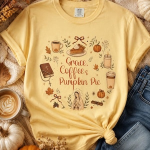 Può includere: Una t-shirt a maniche corte color giallo pallido con una grafica circolare e il testo "Grace, Coffee & Pumpkin Pie". Il design include illustrazioni di una tazza di caffè, una fetta di torta, una zucca, una candela, una Bibbia con rosario e una figura religiosa.