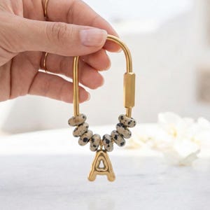 May include: Gold-coloured keychain with a large letter "A" pendant. The keychain features a series of speckled, light-coloured beads. The design includes a curved top and a rectangular bar, creating a stylish and personalised accessory.