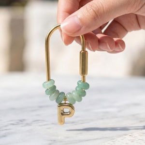 May include: Gold-colored keychain with a jade bead accent and the letter "P". The keychain is held by a hand against a blurred background. The jade beads are light green and the letter "P" is in a serif font.
