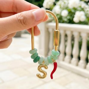 Könnte beinhalten: Goldfarbener Schlüsselanhänger mit einer Kette aus grünen Steinperlen, einem goldenen Buchstaben "S" und einem roten Chili-Pfeffer-Anhänger. Der Hintergrund ist verschwommen und zeigt weiße Blumen und eine weiße Balustrade.