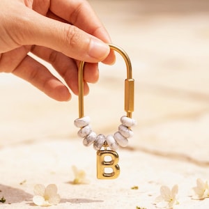 May include: Gold-colored keychain with a white and gray stone bead accent and a gold letter "B" charm. The keychain is held by a hand against a neutral background, with small white flowers scattered around.