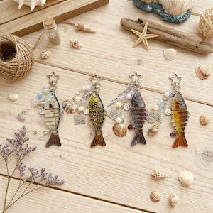 Könnte beinhalten: Vier dekorative Fisch-Schlüsselanhänger mit sternförmigen Verschlüssen, jeweils mit einzigartigem Farbschema und Design. Verziert mit Perlen, Anhängern und Muscheln, liegen sie auf einer hellen Holzoberfläche und erzeugen ein maritimes Thema.