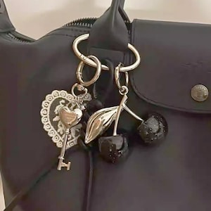May include: Black handbag with decorative keyrings. One keyring has a key and a heart-shaped charm with lace detail. Another keyring features a cherry design with black, glittery cherries and silver leaves. The bag has a top handle and a zip closure.