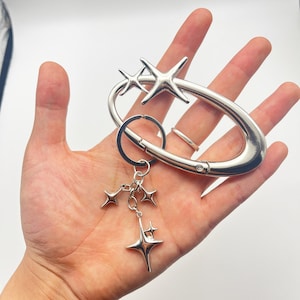 Puede incluir: Un llavero plateado con un mosquetón ovalado grande con dos estrellas. Un grupo de pequeños dijes de estrella cuelgan debajo. El llavero se sostiene en una mano sobre un fondo blanco.