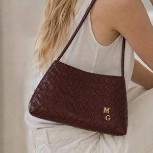 May include: A dark burgundy woven leather shoulder bag featuring a rectangular silhouette and a single thin strap. The front of the bag is adorned with two gold-coloured metal letters, M and G, positioned in the lower right corner of the weave.