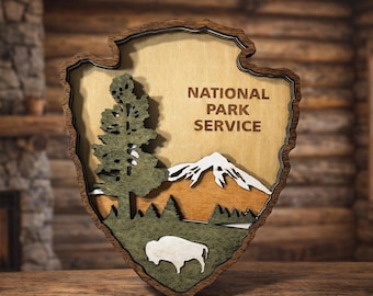 May include: A layered wooden wall plaque shaped like the National Park Service arrowhead emblem. It features a dark brown border, a light wood background with the text NATIONAL PARK SERVICE, a green tree, white mountain peaks, and a white bison.
