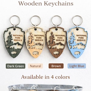 May include: Wooden keychains featuring the National Park Service logo in dark green, natural, brown, and light blue. Each keychain has a metal ring and a shield shape with a mountain and tree design. Available in 4 colors.