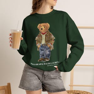 May include: A dark green long-sleeve sweatshirt featuring a graphic of a teddy bear wearing a tan fringed jacket, a red bandana, and blue jeans. The text "POLO BEAR by RALPH LAUREN" is printed in white below the bear graphic on the front of the garment.