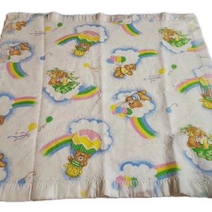 May include: A square white baby blanket featuring a repeating pattern of brown teddy bears in hot air balloons, floating on clouds, and riding rainbows. The fabric is decorated with colorful balloons and has a ruffled border trim.