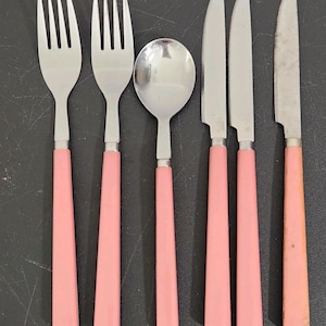 May include: A set of six vintage stainless steel flatware pieces: two forks, a spoon, and three knives. Each piece has a pink handle. The flatware is arranged on a dark surface.