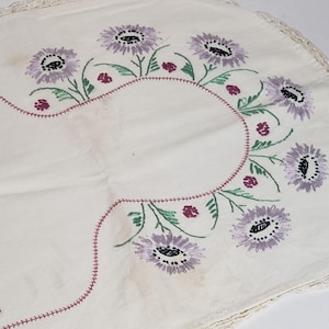 May include: Cream-colored embroidered tablecloth featuring a floral design. The flowers are lavender with black centers, green stems, and small maroon accents. The edge has a delicate lace trim.