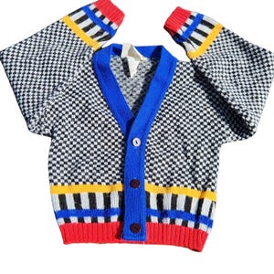 May include: A vintage cardigan sweater with a black and white checkered pattern. The sweater has a blue trim, red cuffs, and yellow and black stripes. It features three brown buttons and a V-neck.