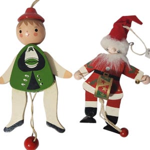 May include: Two wooden Christmas ornaments. One is a figure in a green vest and red hat. The other is a Santa Claus figure in a red suit and hat. Both have dangling pull strings and are painted with festive details.
