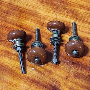 May include: Four brown ceramic cabinet knobs with a smooth, glossy finish. Each knob features a rounded top, a metallic base, and a threaded metal bolt extending from the bottom, accompanied by a matching metal washer and a hexagonal nut for installation.