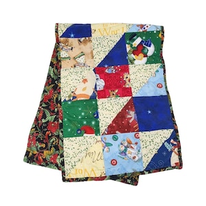 May include: A patchwork quilt with a Christmas theme. The quilt features squares and triangles in various colors, including red, green, blue, and cream. The quilt has images of snowmen, angels, and reindeer. The border is a black fabric with red and green floral patterns.