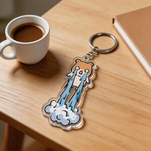 May include: A clear acrylic keychain featuring a cartoon bear with tears streaming down, forming a cloud. The bear is light brown with a white belly. The keychain has a silver metal ring. A cup of coffee and a notebook are in the background.
