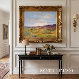 May include: An ornate gold framed landscape painting hangs above a black console table. A lamp, books, and a vase of dried flowers sit on the table. The wall is white with decorative molding. The text "SMITH & IMAGO THE HEIRLOOM COLLECTION" is at the bottom.