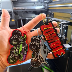 May include: Three acrylic keychains with ball chains. One features a green and black monster truck with a red border, another a grey shark-themed monster truck with a blue chain, and the third a red and black sign with the text ZOMBIE.