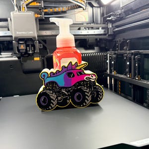 May include: A red plastic soap dispenser with a white pump mechanism, partially obscured by a colorful 3D-printed unicorn monster truck figure featuring a pink, blue, and purple body with large black tires and a yellow horn, resting on a flat surface.