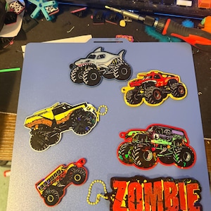 May include: A collection of six colorful monster truck keychains and one rectangular keychain with the word ZOMBIE in bold red and yellow letters. The designs include a shark-themed truck, a red truck, a yellow truck, and a green truck.