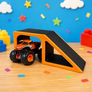 May include: An orange monster truck toy with black wheels and a cartoon face, positioned in front of an orange and black ramp. The background features a blue sky with star and cloud decorations, and colorful confetti.