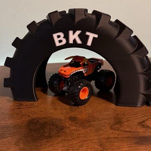 May include: A black, arched tire-shaped display stand with the white text BKT printed on the top. Positioned underneath the arch is a small orange monster truck toy featuring black tires with bright orange rims and a bull-themed graphic design.