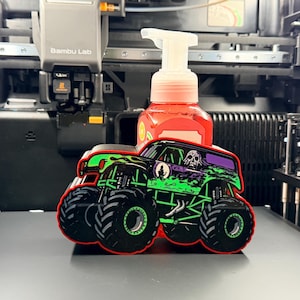 May include: A red liquid soap dispenser featuring a custom 3D-printed monster truck holder. The holder is black with bright green flame graphics, large black tires with green rims, and a skull illustration on the side of the truck body.