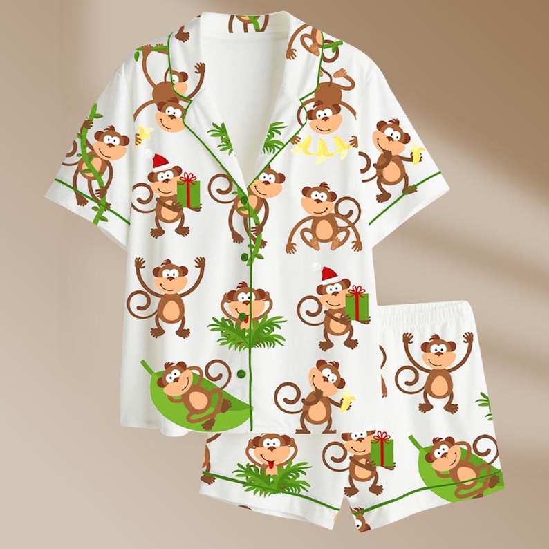 May include: White pajama set with a repeating pattern of cartoon monkeys in various poses. The short-sleeved top and shorts have green trim and accents. Some monkeys wear Santa hats and hold gifts.