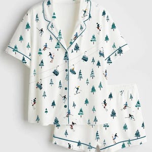 May include: White pyjama set with a ski resort design. The short-sleeved button-up shirt and matching shorts feature a repeating pattern of skiers, pine trees, and ski lifts in teal and black. The edges of the set are trimmed with teal piping.